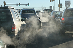 Cars sit at a stoplight with visible pollution from the tailpipe emissions.